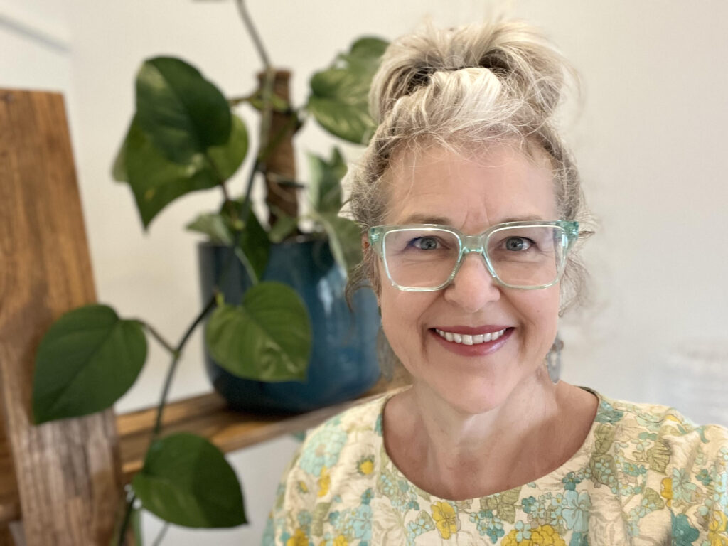 woman with hair in a top know with a smmile, wearing blue glasses and a floral top infront of a plant